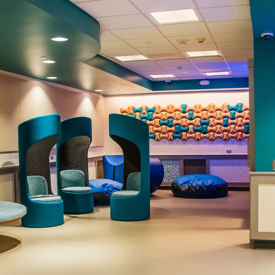 Ten obraz przedstawia nowoczesne i stylowe wnętrze z zakrzywionymi niebieskimi krzesłami i artystycznymi elementami na ścianie. Każde krzesło ma okrągły, jasnoniebieski podnóżek i zapewnia prywatność dla użytkowników. Na ścianie w tle znajduje się artystyczna instalacja składająca się z wielu kolorowych kształtów tworzących efekt 3D. Oświetlenie jest miękkie i nastrojowe, co sprzyja relaksowi.