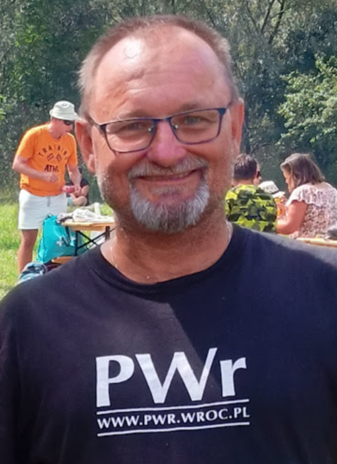 Łysiejący, uśmiechnięty mężczyzna w średnim wieku. Nosi okulary i czarną koszulkę z napisem PWr