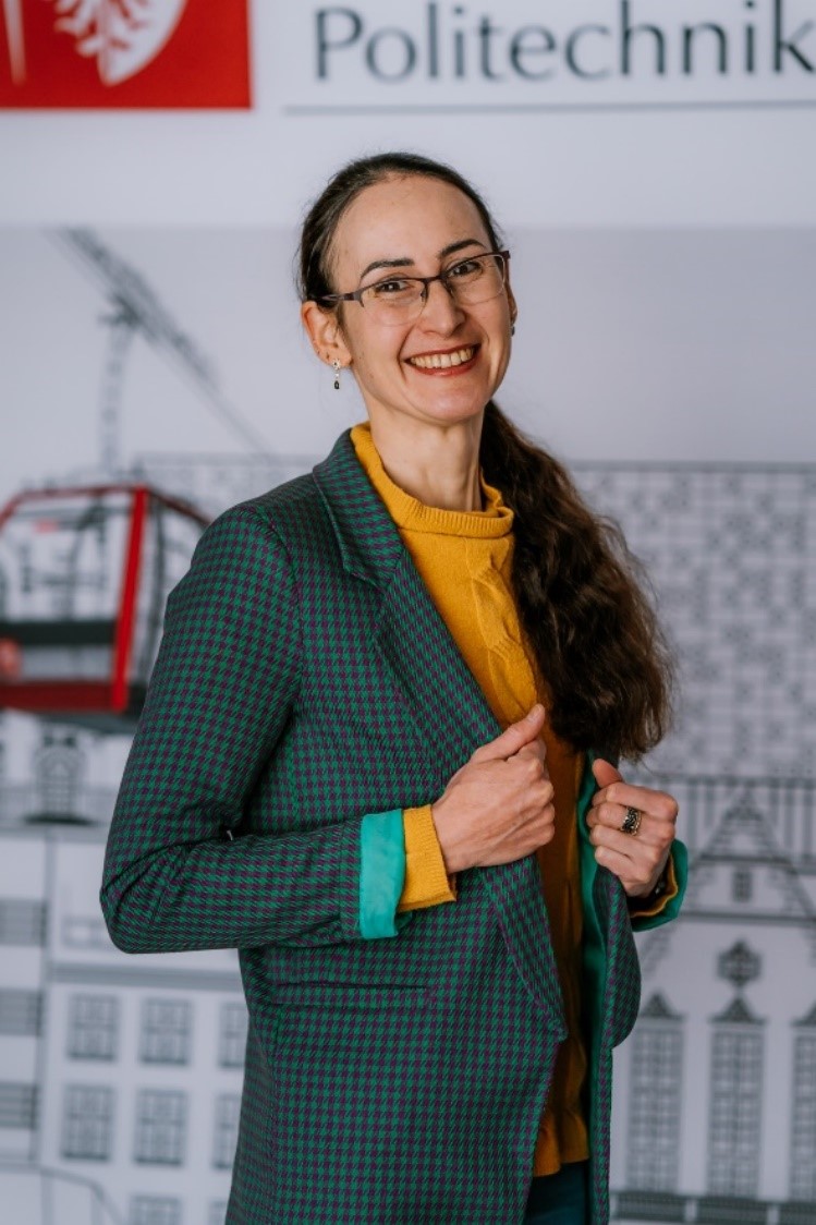 Katarzyna Helios standing. Smiling radiantly. Long, dark hair and glasses.