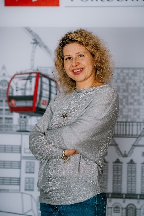 A woman with a smile on her face, she is medium build, with shoulder-length curly hair. In the background, the cable car of the Wrocław University of Technology.