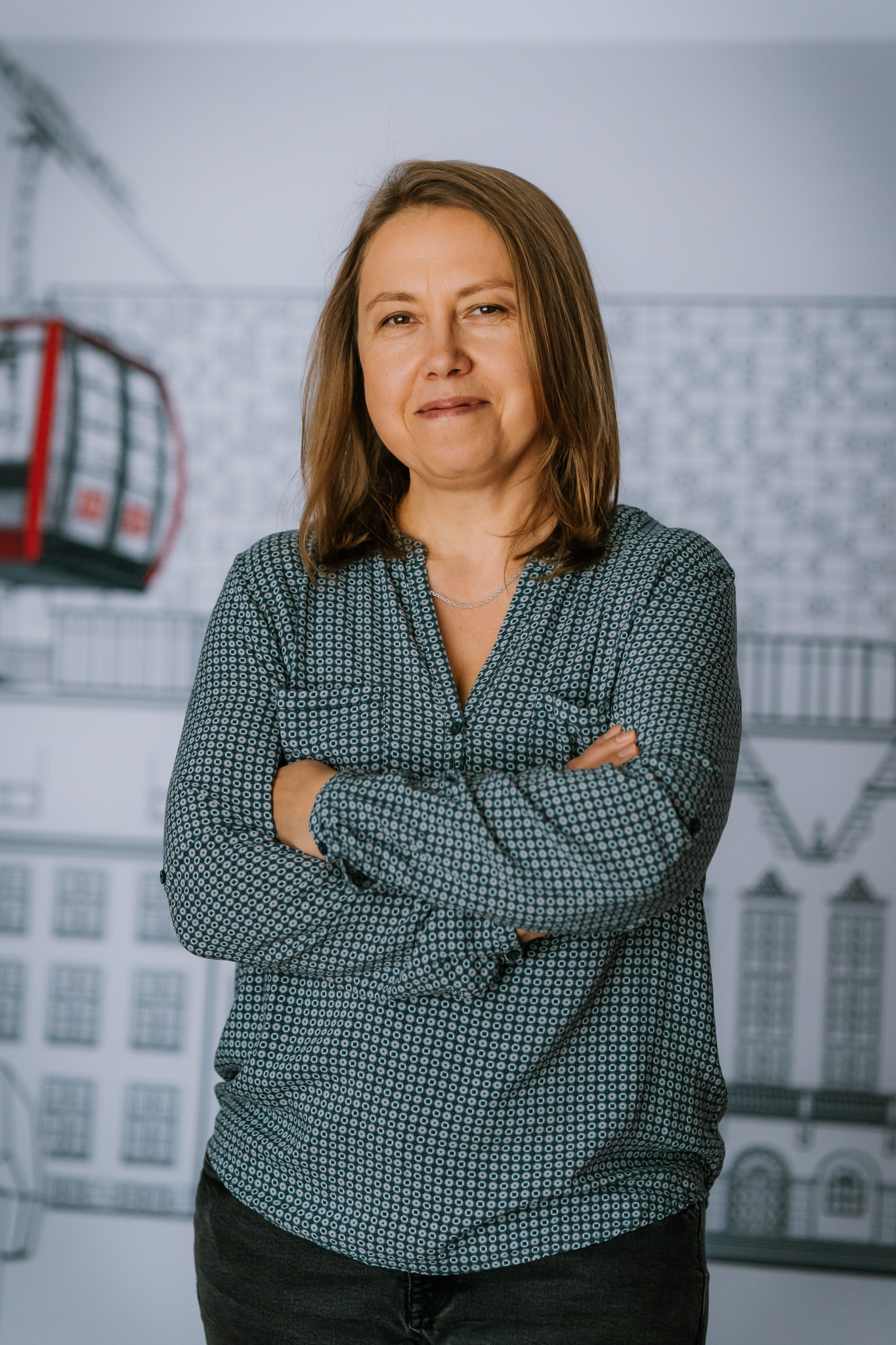 A woman stands with her arms folded in front of her and smiles in a friendly manner. She has light, straight, medium-length hair. In the background: a drawing of the main building of Wrocław University of Science and Technology with a red Polinka cable car.