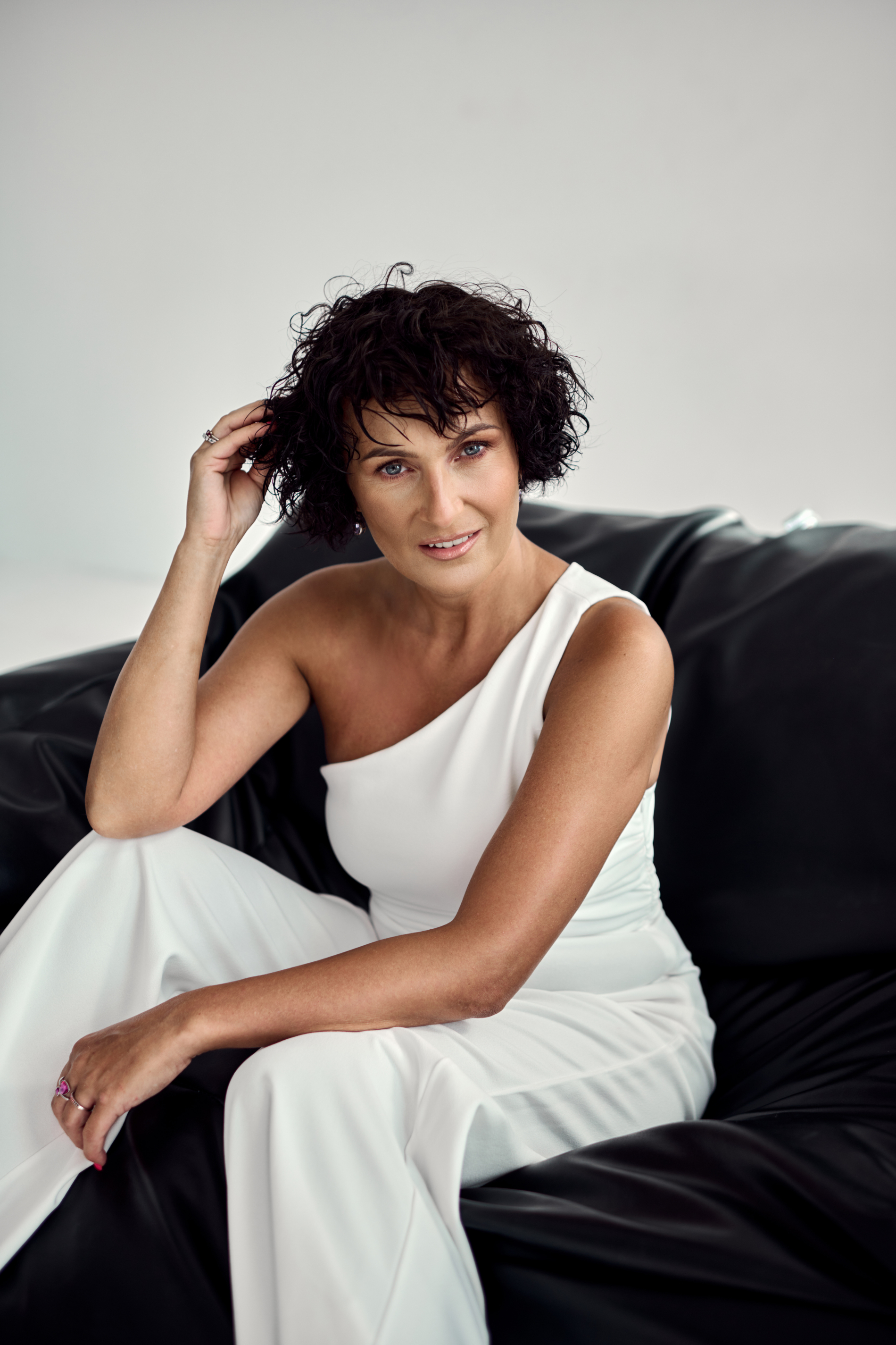 Photo with alt text: smiling woman in a black jumper, short, dark hair, hands clasped on her stomach, a lot of colourful jewellery. Bust with round windows in the background.