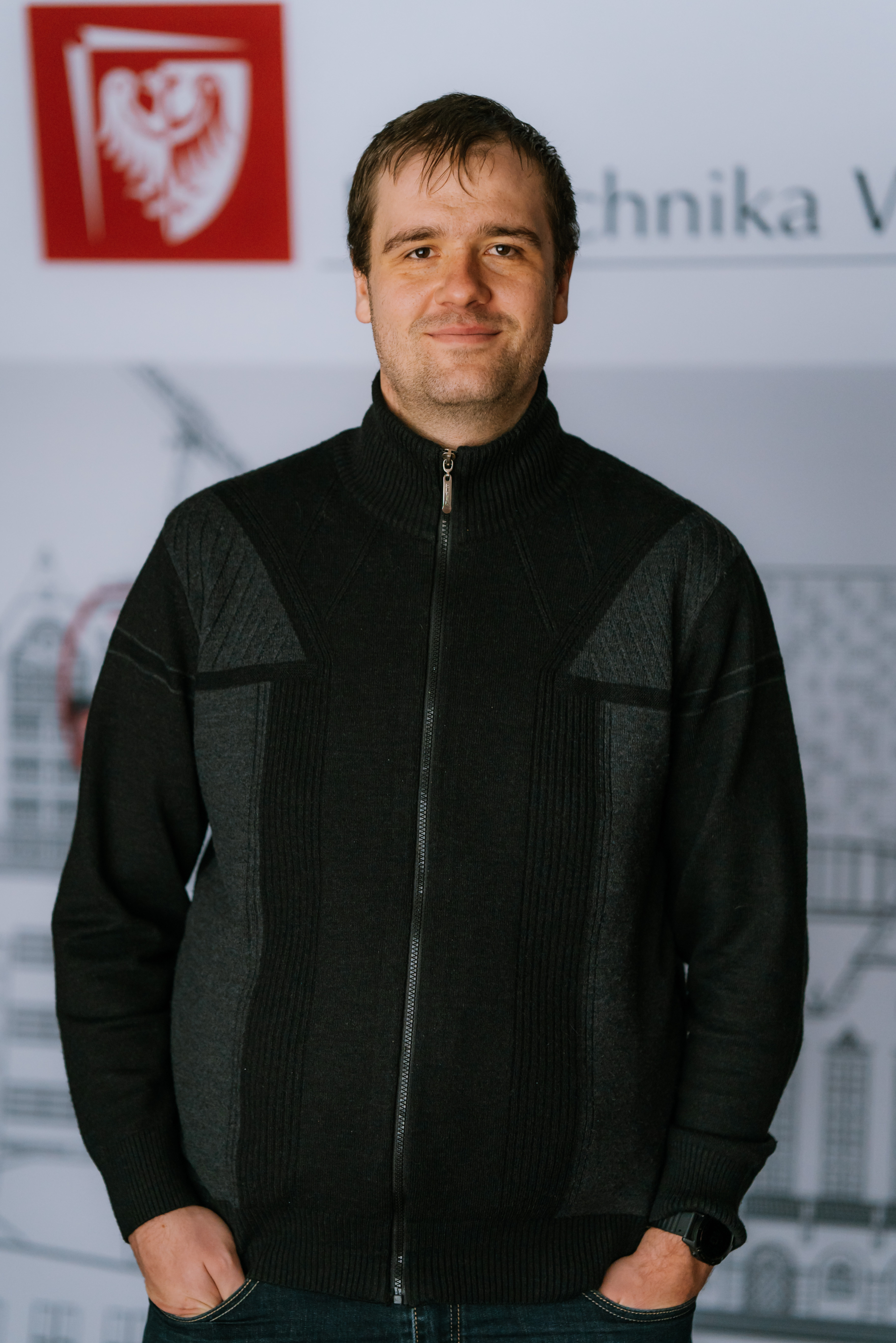 Tall man, 30 years old. He is standing in the front of a banner with the logo of the Wrocław University of Science and Technology. He is wearing a dark sweater fastened with a zipper, he has his hands in his pants pockets and smiles softly.